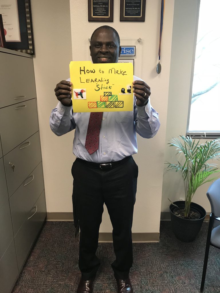 A man holds a homemade sign that reads, "How to Make Learning Stick."