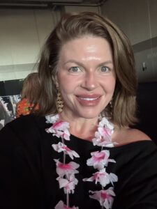 Close-up selfie of a smiling woman with shoulder-length brown hair, wearing a black off-the-shoulder top, gold dangle earrings, and a pink-and-white paper flower lei, standing indoors.