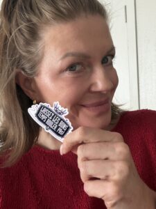 Close-up selfie of a smiling woman with brown hair pulled back, wearing a red sweater, holding a purple-and-white sticker that reads "Laissez les bons temps rouler, PAs!" near her face.