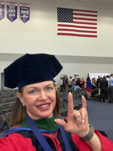 A selfie of a smiling woman in red and green doctoral regalia with a black velvet tam, making an "I love you" sign in ASL, with an American flag in the background.